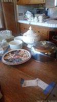 Photo showing white ceramic tureen with lid, two white bowls, floral patterned plates, and silver-tone cake cover arranged on wooden table.