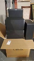 Six Carver cinema speakers stacked on a cardboard box with visible Carver logo. Speakers include subwoofer, center, satellite, and main units.