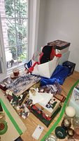Wide view of the collection of holiday items stacked on a wooden floor near window, showing fabric tree skirt, boxes, ribbons, and snowman figure