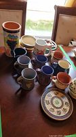 Overview of ceramic set including coffee mugs, bowls, pitcher, wine cooler, and plates all placed on wooden table