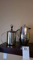 Photo shows two stainless steel coffee presses side by side on a dark surface against a neutral background.