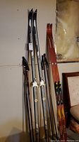Two sets of cross country skis standing against a wall. One pair is black and white with step-in bindings, approximately 210 cm long. The other pair is red and yellow with toe clip bindings, approximately 195 cm long. Several poles are visible leaning with skis.