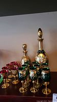 Two Bohemian handpainted sets including decanters and glasses; green and red glasses with floral gold decoration visible