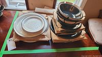 Two stacks of dinnerware and bowls on cardboard. Left stack consists of white Royal Doulton plates with silver rims and embossed floral center pattern. Right stack has white bowls with slight black tint inside.