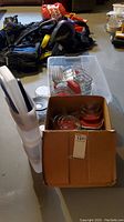 Overall view showing a cardboard box with multiple glass food storage containers with red lids, some metal rack containers, and a stack of white and blue plastic storage containers.