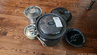 Overview of all cookware items on wooden floor: two stainless steel pots with lids, black speckled roasting pan with lid, Circulon frying pan with lid.