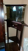 Photo showing wooden corner side table with single drawer and lower shelf, dark polished finish, placed in corner beside a chair.