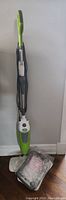 Full upright view of steam mop with handle, cord wrapped around hooks, and mop head wrapped in plastic on floor.