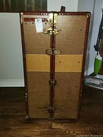 Front exterior view of the standing MendelTrunx steamer trunk showing fabric panels, leather trim, brass corner reinforcements, and locking clasps.