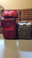 Photo showing large red upright suitcase with small matching red travel bag on top and medium green upright suitcase next to them on floor in front of plaid couch.