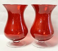 Pair of ruby red glass vases with flared mouths and hourglass shapes on clear pedestal bases, side view.