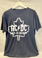 Front view of black AC/DC concert T-shirt size large featuring white maple leaf and 'AC/DC BACK IN CANADA' text.