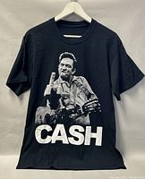 Front view of black T-shirt showing large grayscale graphic of Johnny Cash holding guitar and showing middle finger with 'CASH' text below
