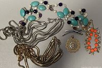 Lot includes silver and gold-toned necklaces, a gold-tone locket, and orange stone bracelet all laid out on white surface.