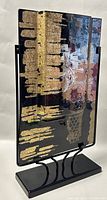 Rectangular fused glass vase with gold leaf and black abstract designs, mounted on a black curved metal stand, photographed from front angle.