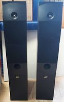 Pair of black Energy L-26 tower speakers front view with grills on showing tweeters and port holes near the base, standing upright on floor.