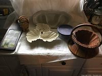 Photo of assorted kitchenware and collectibles on white cloth including candy dish, wooden basket, Pyrex dish, tin box, and small container.