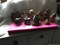 Photo showing the full set of 9 ceramic village pieces spaced on a pink mat, with visible snow detail accents and varying architectural styles.