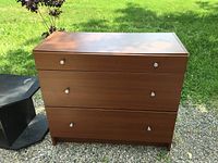 Front view of the three-drawer wooden dresser with white knobs, medium brown finish, placed outdoors.