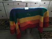Crocheted rainbow color blanket draped over a small table in a kitchen setting, showing its multi-color stripes and texture.