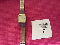 Front view of vintage wristwatch showing gold-tone rectangular dial and metal link bracelet on pink background with lot number card.