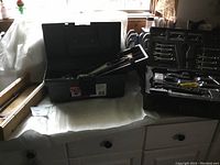 Photo of one open black toolbox with hand tools visible inside including wrenches, sockets, screwdriver bits and other hand tools on white cloth-covered surface.