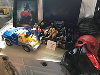 Overall view of toys and costumes arranged on a white table, featuring the blue tow truck, action figures, black car, red helmet, and black cape.