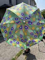 Photo of one beach umbrella opened showing full floral seashell pattern on green fabric, on pavement outdoors.