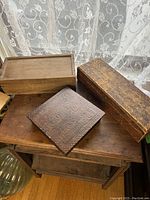 Three wooden boxes displayed together on a wooden surface by lace curtains. One box has a carved floral lid pattern.