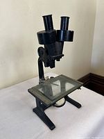 Full view of the antique microscope showing binocular eyepieces, glass stage with metal stage clips, and sturdy metal base.