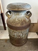 Front view of antique metal milk can with handles and painted lettering 'GUELPH CRY' on body