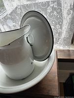 Photo showing the white enamelled ironstone pitcher resting inside the wash basin, both having black trim around rims.