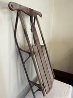 Side view of antique wooden sleigh showing metal frame and runners, wooden slat deck, and curved handlebar.