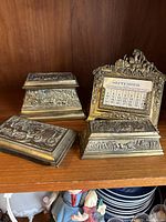 Four brass desk items displayed on a wooden shelf including a perpetual calendar, ink well, paperweight, and a small box.