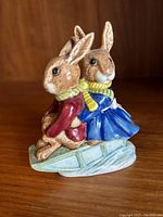 Front and side view of the Bunnykins sleigh ride figurine showing two rabbits back-to-back on a sleigh, dressed in winter outfits with scarves.