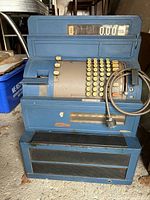 Front view of blue mid century modern National Cash Register (NCR), showing cream keys, mechanical numeric display, receipt compartment, cash drawer, and attached power cord.