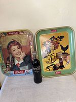Photo showing two Coca-Cola trays and one unopened glass bottle placed between them against a plain wall.