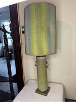 Table lamp with ceramic green glaze cylindrical base, metal ring accents, square metal bottom, and tall drum lampshade with vertical stripes in blue, green, and yellow fabric.