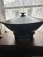 Side view of black Mid Century Modern electric wok with lid and knob handle, set on wooden surface with lace curtain background.