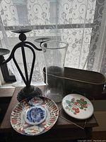 Photo showing the glass vase, metal candleholder, two decorative plates, and the metal basket on a wooden surface by a window with lace curtains.