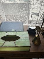 A brass-tone decorative bell with handle, next to a green and pink stained glass tissue box cover on a wooden surface, near a window with lace curtain.