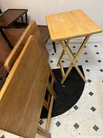 Three Mid Century Modern wooden TV trays, two folded and leaned against a wall and one standing upright on a black rug on a tiled floor.