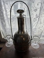 Silver-tone metal decanter with stopper and handle alongside two clear glass goblets with etched decoration, placed in front of a lace curtain.