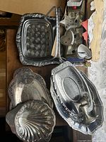 Photo of multiple silver trays stacked and arranged on a wooden surface, showing the variety of sizes and edge designs including shell-shaped and ornate patterns.