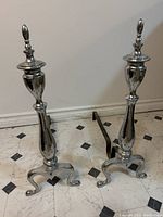Pair of vintage silver-finished fireplace andirons in a standing position showing full body shape including the curved tripod base and pointed finials.