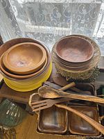 Various sized round wooden bowls stacked together, rectangular woven serving tray with wooden compartments, long wooden spoon and fork resting on tray, gold-green fabric hot pads beside them.