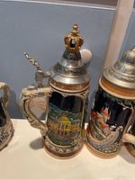 Photo of beer stein with a golden crown-shaped pewter lid and colorful architectural scene on ceramic body.