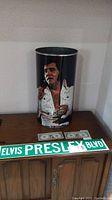 Metal can featuring color image of Elvis Presley in white jumpsuit with microphone, green 'Elvis Presley Blvd' street sign, and two modified $1 bills with Elvis face on wooden cabinet.