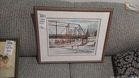 Framed winter landscape print showing a metal bridge over a river with snowy banks, resting on a sofa.