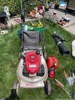Full front view of mower on grass showing deck, engine, handle, fuel can, and grass bag accessory.